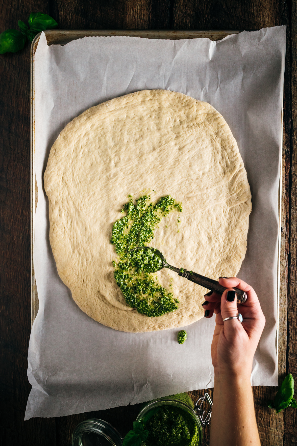 A person using a spatula to spread pesto on a pizza dough.