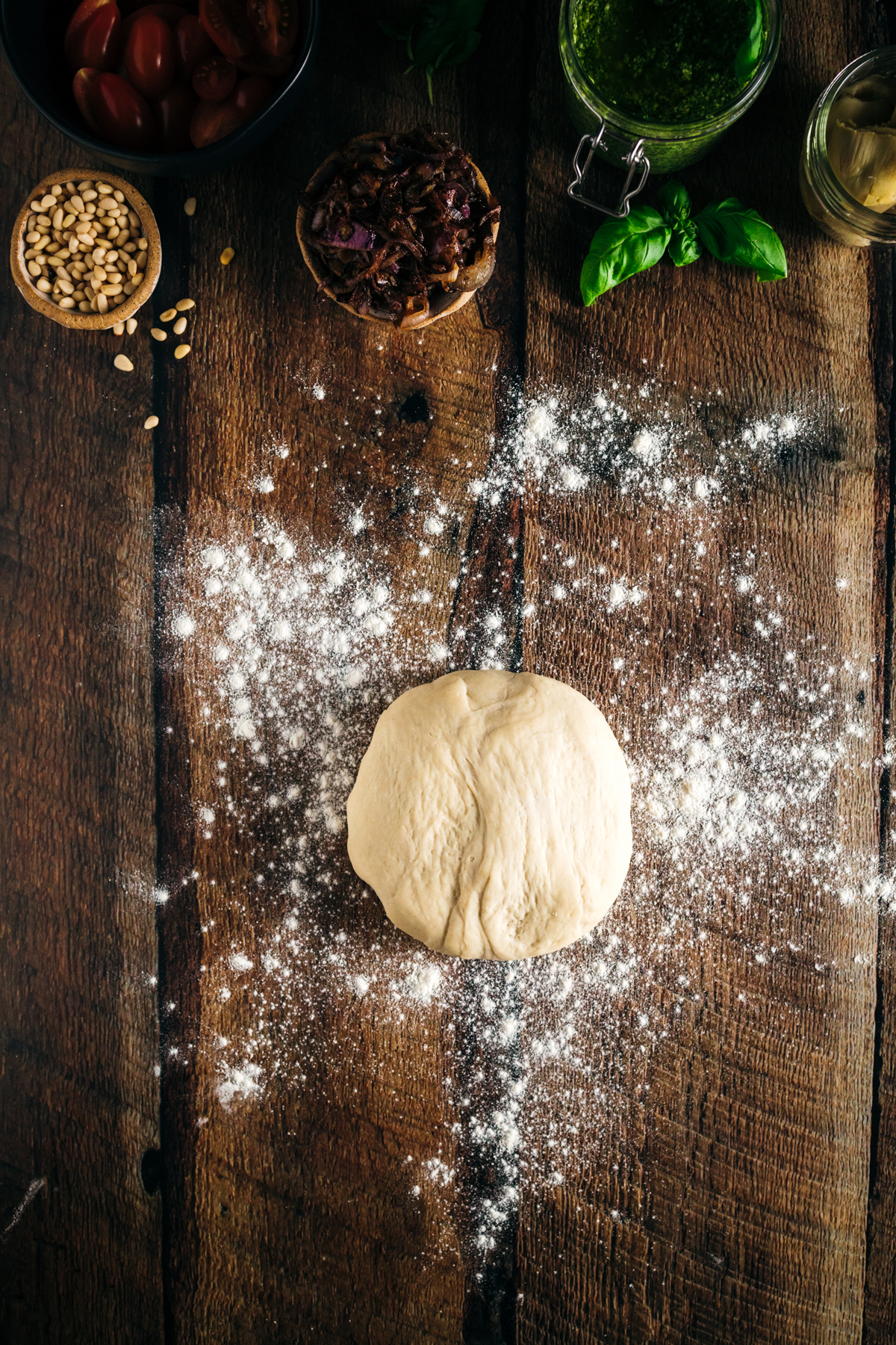 A pizza dough on a wooden table with other ingredients.