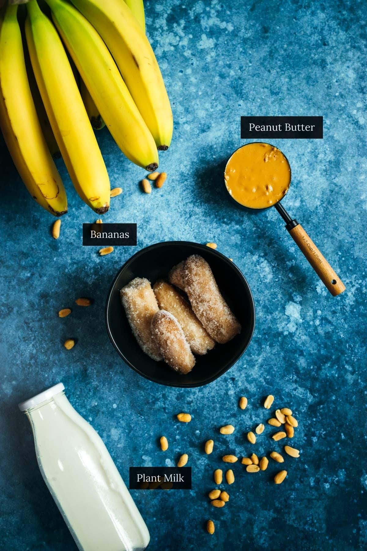 A bunch of bananas, a measuring cup of peanut butter, a bowl of frozen bananas, a bottle of plant milk, and peanuts are artfully displayed on a blue surface.