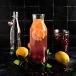 Glass pitcher with pink pomegranate water, lemon slices, and scattered pomegranate seeds, surrounded by whole lemons, fresh mint leaves.