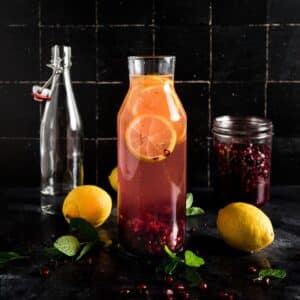 Glass pitcher with pink pomegranate water, lemon slices, and scattered pomegranate seeds, surrounded by whole lemons, fresh mint leaves.