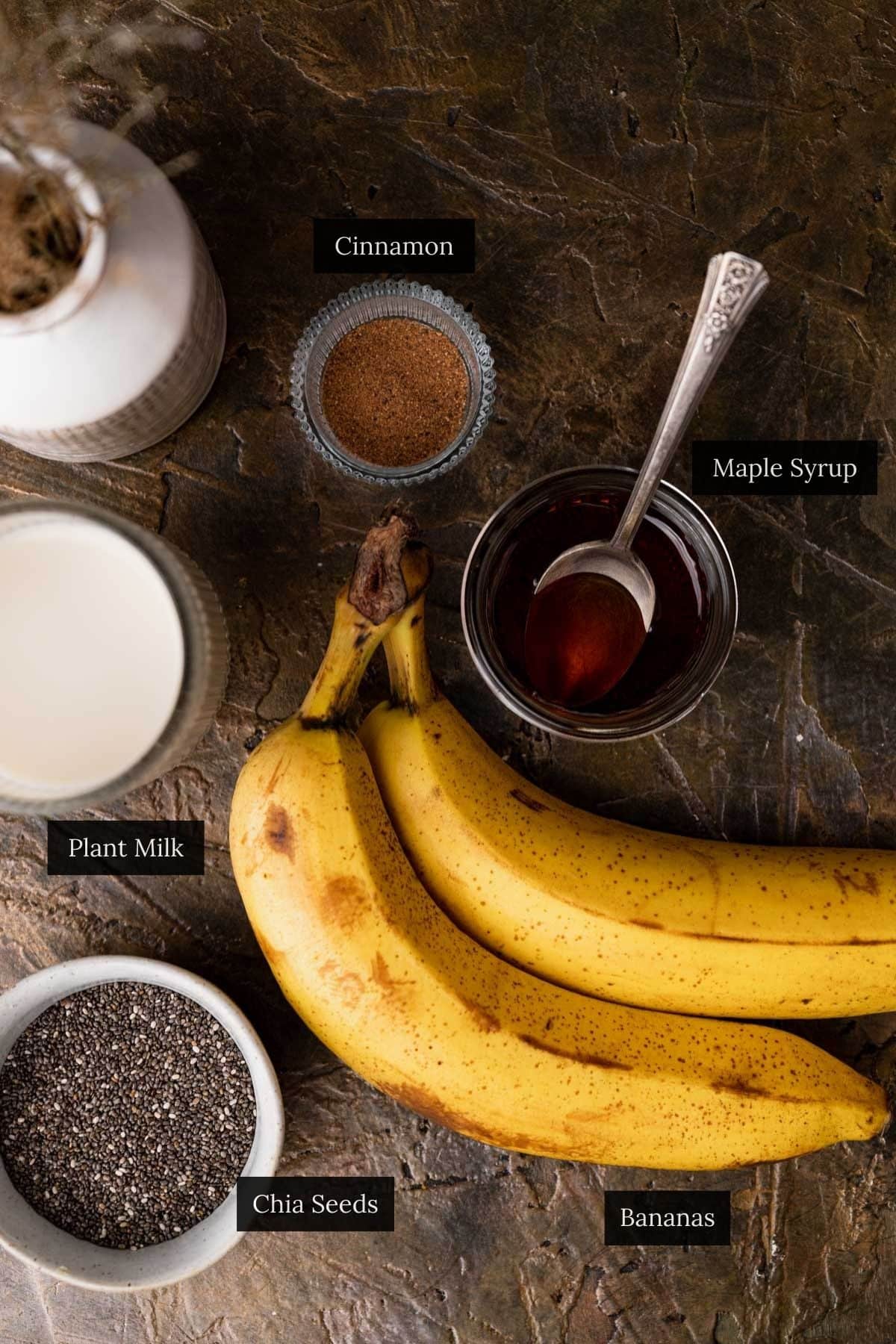 Top-down view of ingredients on a textured surface for banana chia pudding: bananas, chia seeds, plant milk, maple syrup, and a small bowl of cinnamon.