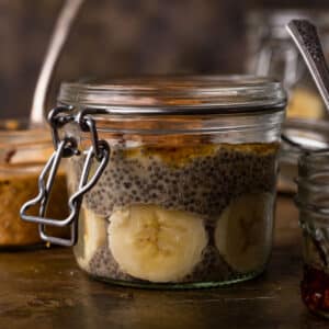 A jar of banana chia pudding with visible slices sits on a rustic surface, surrounded by other jars in the background.