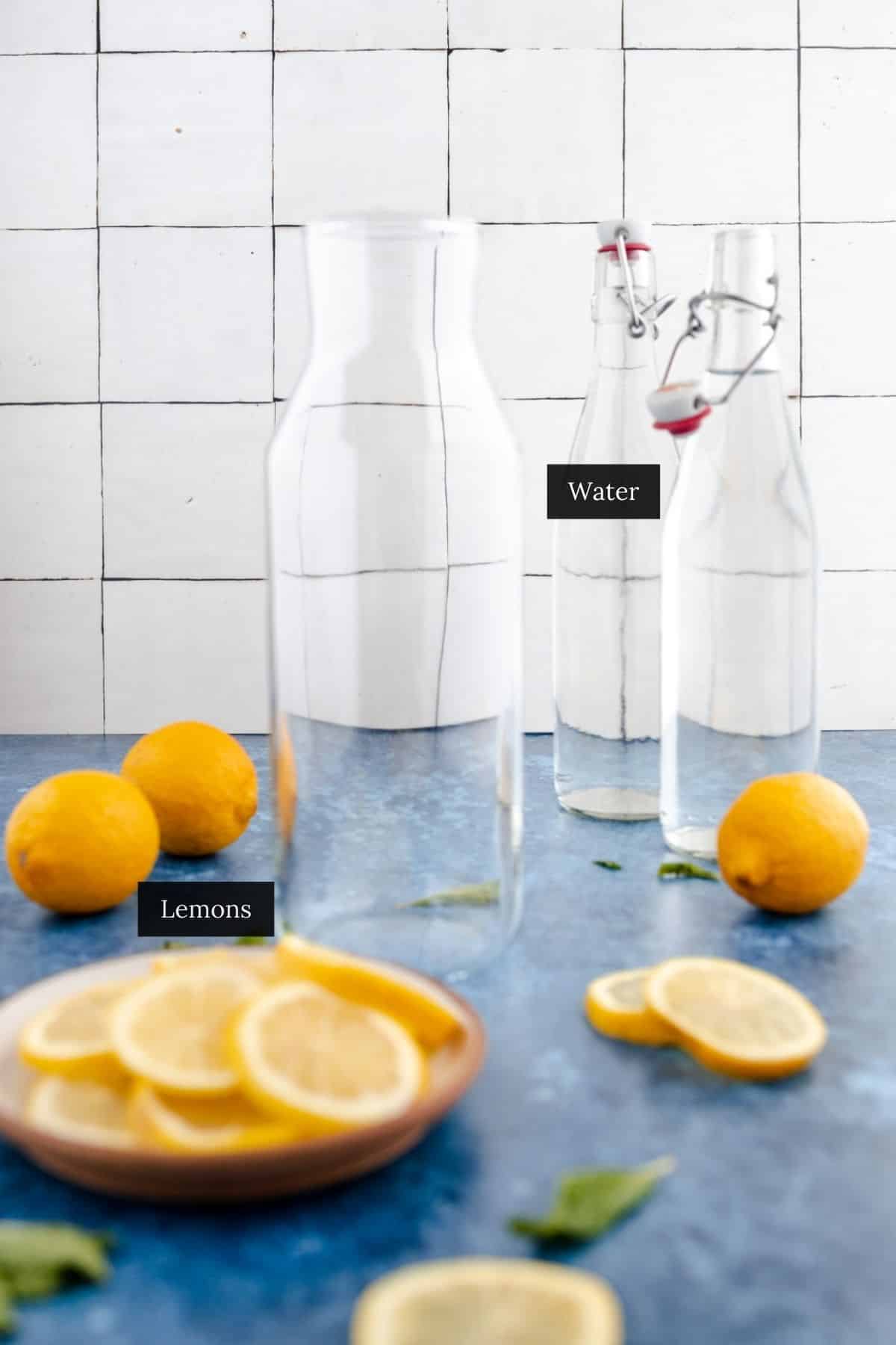 Glass bottles of water and whole lemons are placed on a blue surface with a small dish of lemon slices in the foreground.