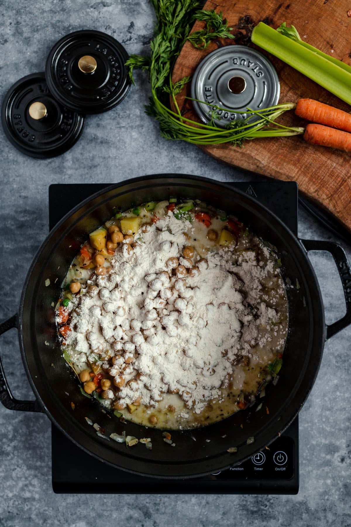 A pot of vegetable stew with flour being added, on a stove with ingredients around it.