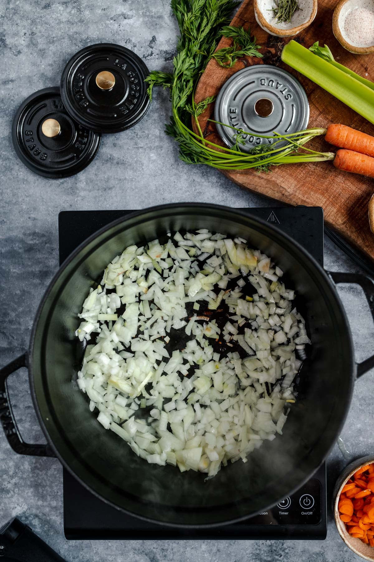 Diced onions being sautéed on a modern stove with various vegetables and herbs prepared on the side.