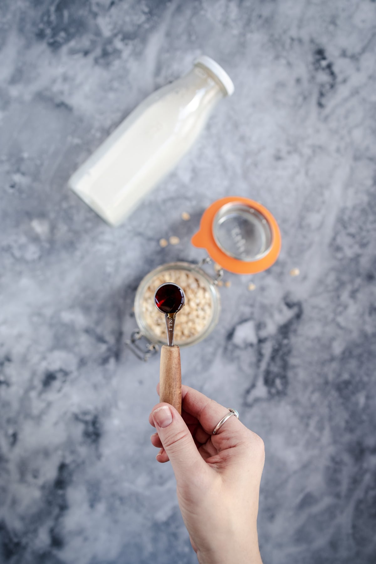 A person pouring syrup into a bowl of oatmeal next to a bottle of milk on a marble surface.