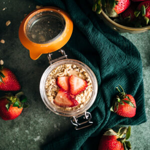 Overhead view of a glass jar filled with oatmeal and fresh strawberries on a dark green cloth, with whole strawberries scattered around.