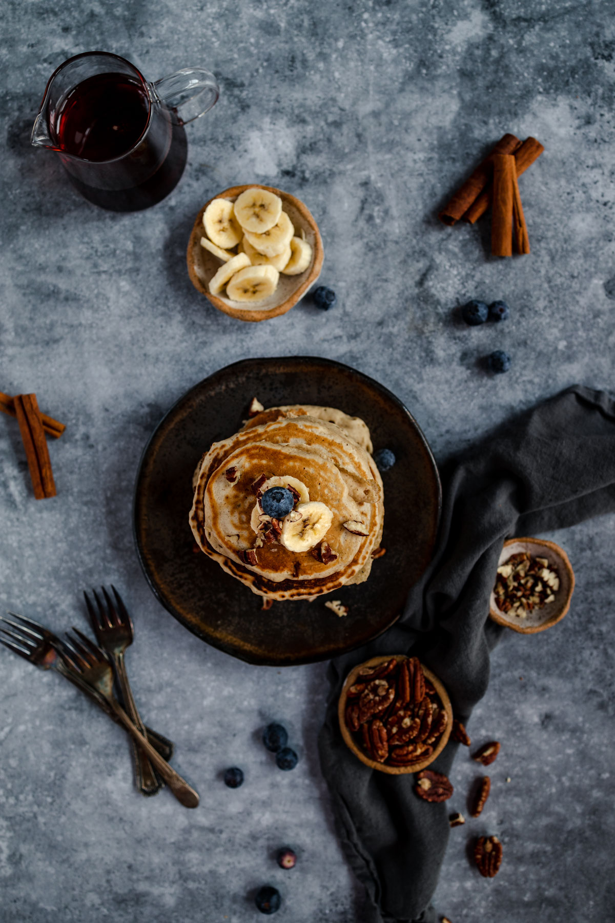 A stack of pancakes with banana slices and a blueberry on top, accompanied by a jar of syrup and scattered nuts and berries on a textured surface.