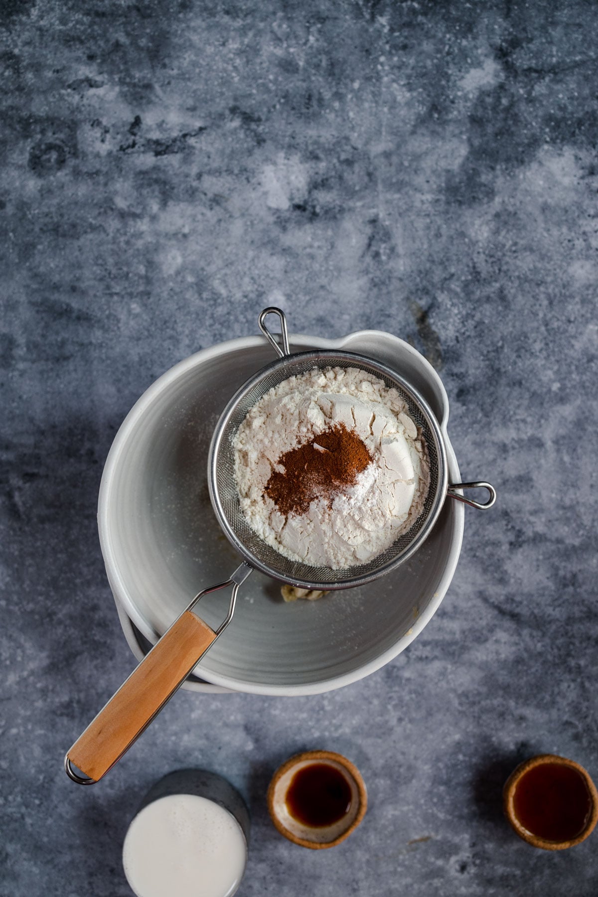 Flour and cinnamon in a sieve over a mixing bowl on a marbled surface with ingredients nearby.