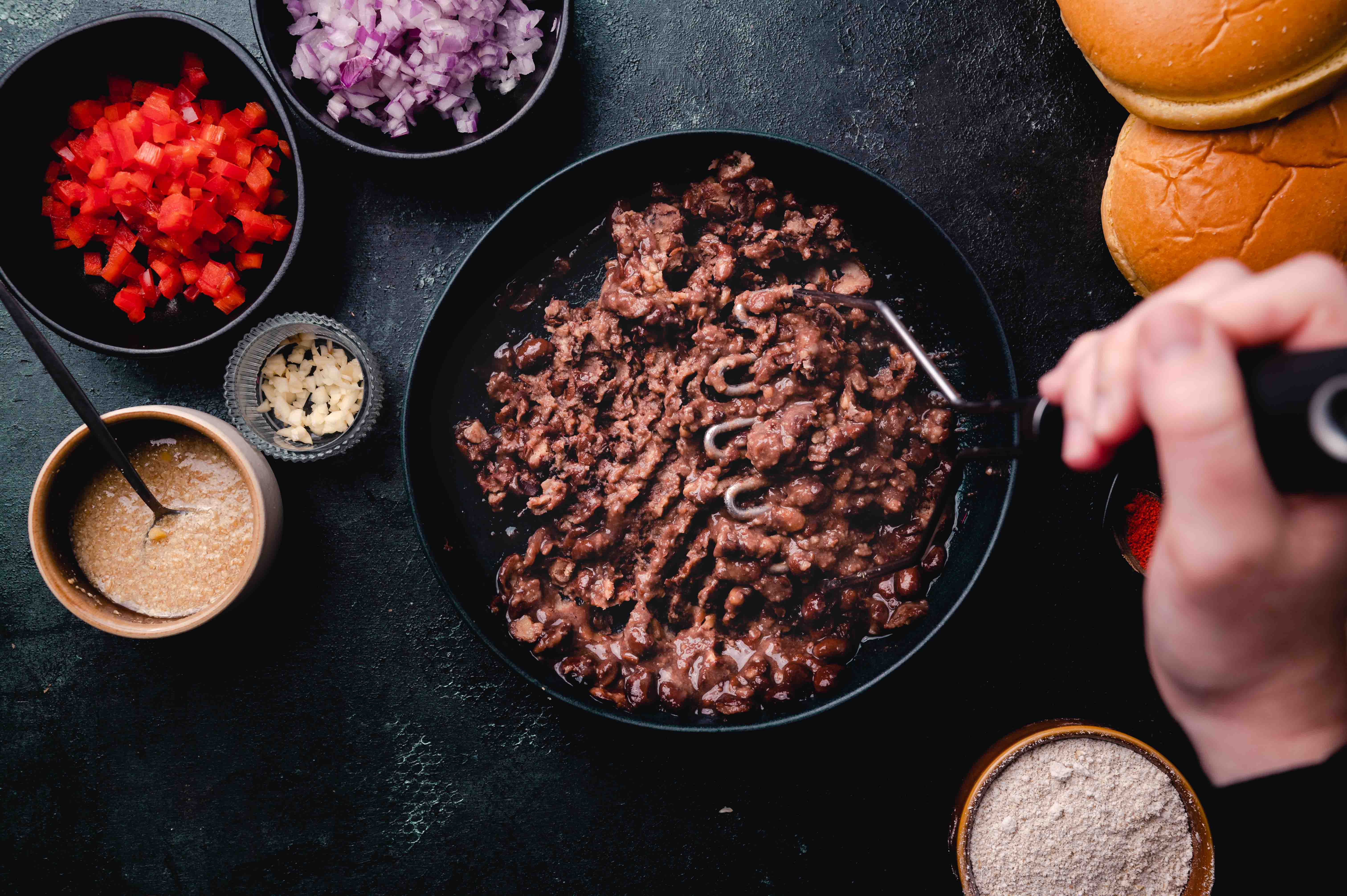 Cooking ingredients for burgers arranged on a dark table, including a pan of ground meat, chopped vegetables, buns, and sauces.