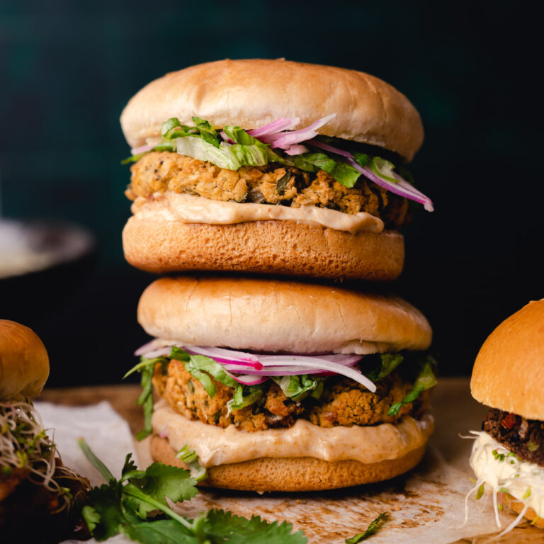 Two stacked chickpea burgers with red onion and cilantro on sesame buns, presented on a wooden surface with moody lighting.