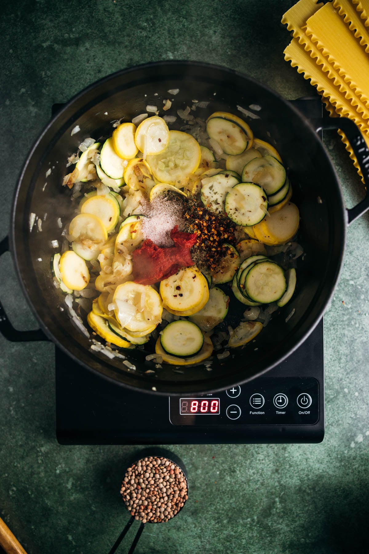 Sliced zucchini and onions cooking in an electric skillet with seasoning, displayed next to uncooked pasta and lentils.