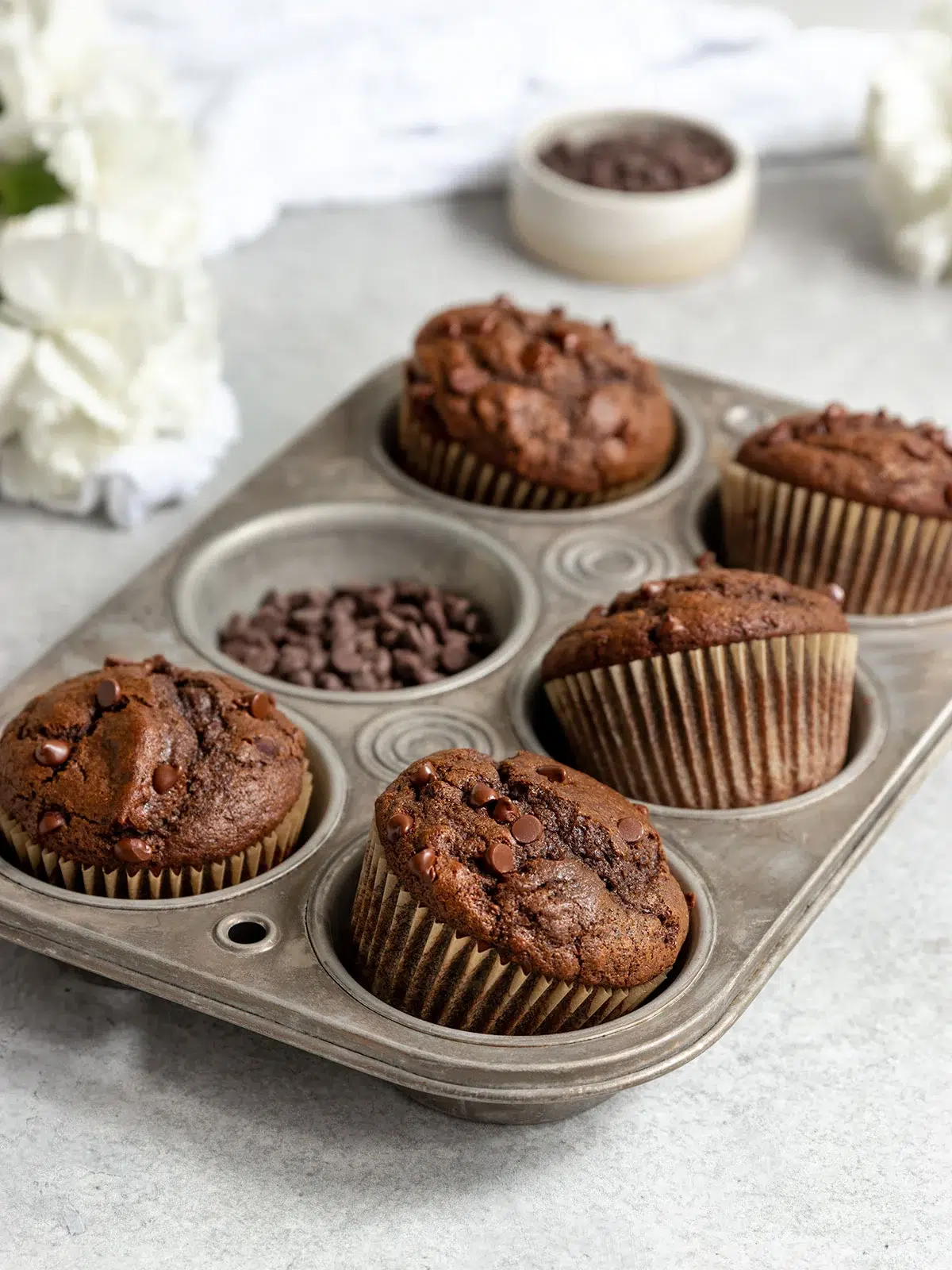 Six vegan chocolate muffins with chocolate chips in a metal baking tray on a marble surface, with flowers and more chips nearby.