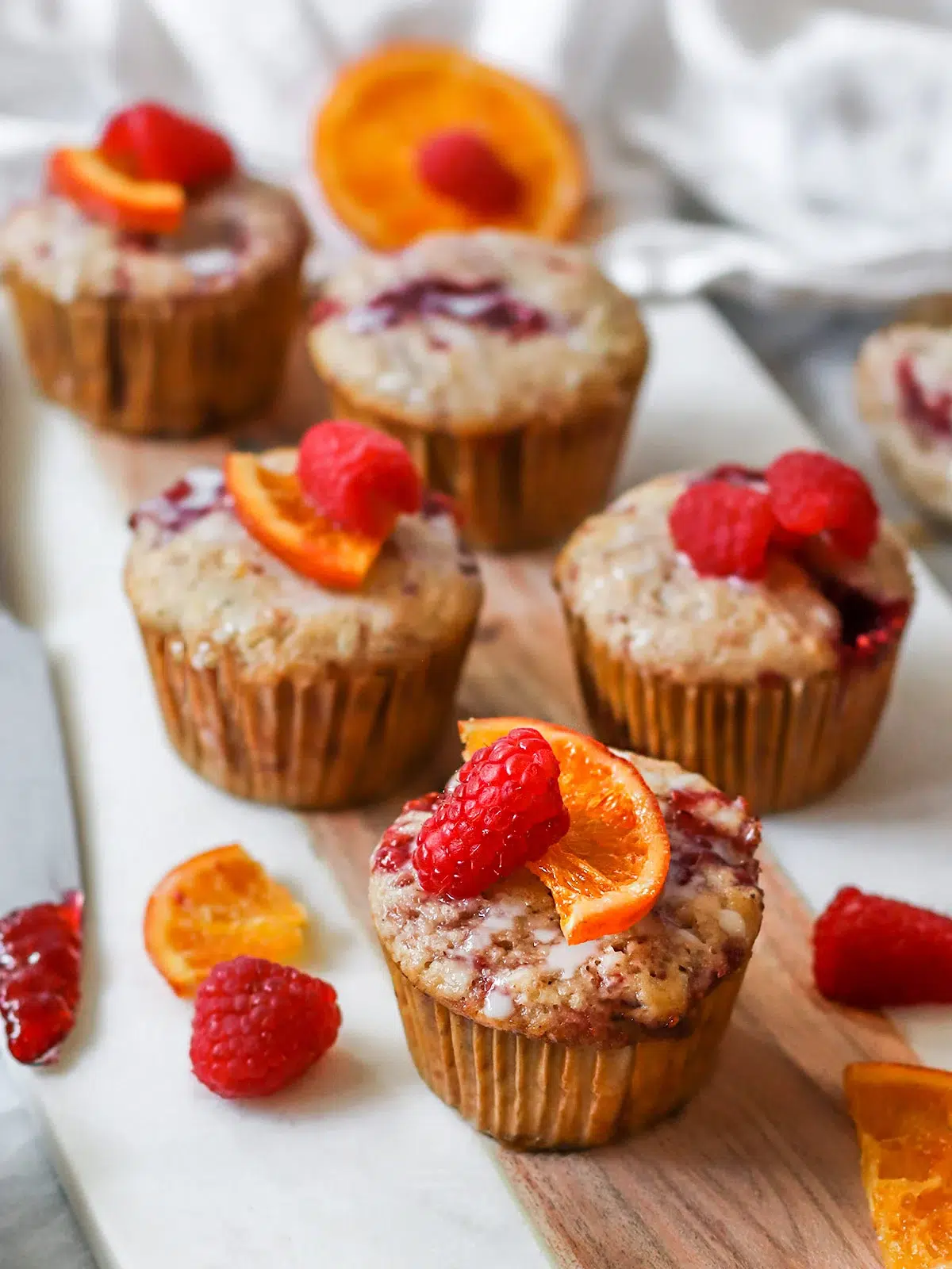 An array of freshly baked vegan muffins topped with raspberries and orange slices, displayed on a marble countertop.