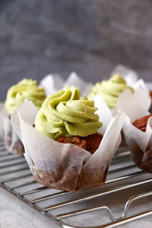 Three vegan carrot muffins with green matcha frosting, served in white parchment wrappers on a wire rack.
