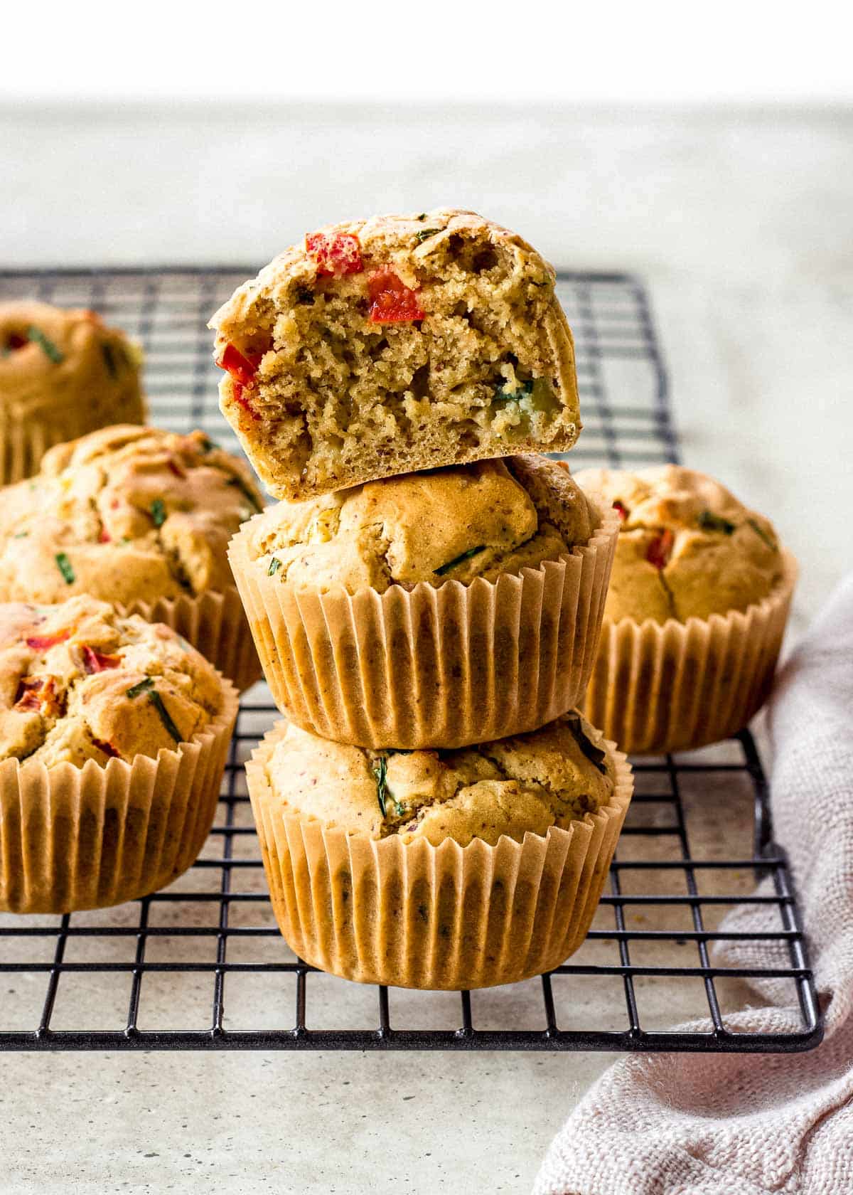A stack of savory vegan muffins with visible chunks of red and green vegetables on a cooling rack, with a bite taken out of the top muffin.