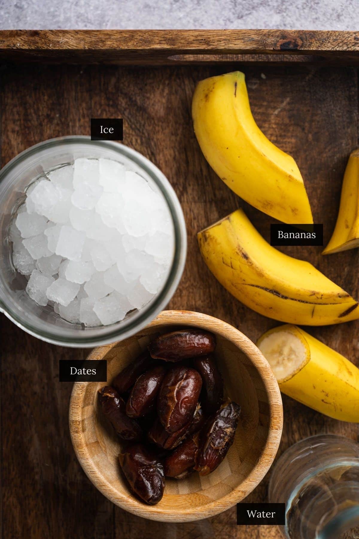 A bowl of ice, bananas, a bowl of dates, and a glass of water on a wooden tray.