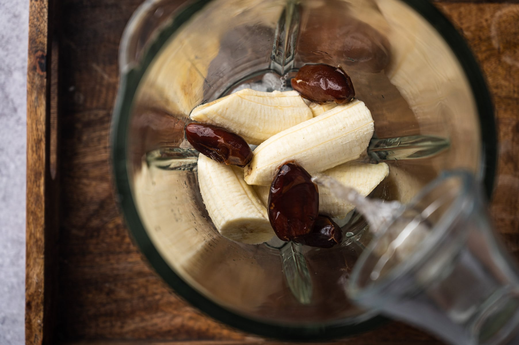 A blender jar with cut bananas and dates, being filled with water from a glass jug on a wooden surface.