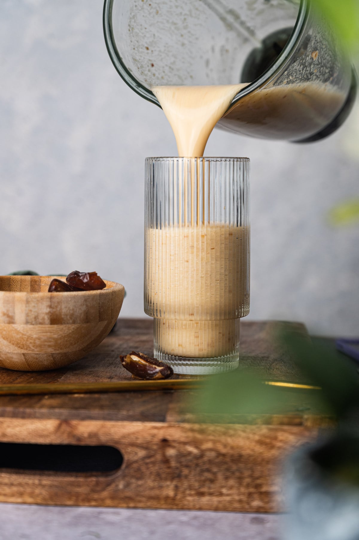 A glass is being filled with a creamy beverage from a blender. A wooden bowl with dates and date pits is on the left, resting on a wooden surface.