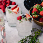 Two glasses filled with ice, garnished with strawberries, raspberries, blueberries, and fresh herbs are placed on a table. Bowls of berries and additional herbs are in the background.