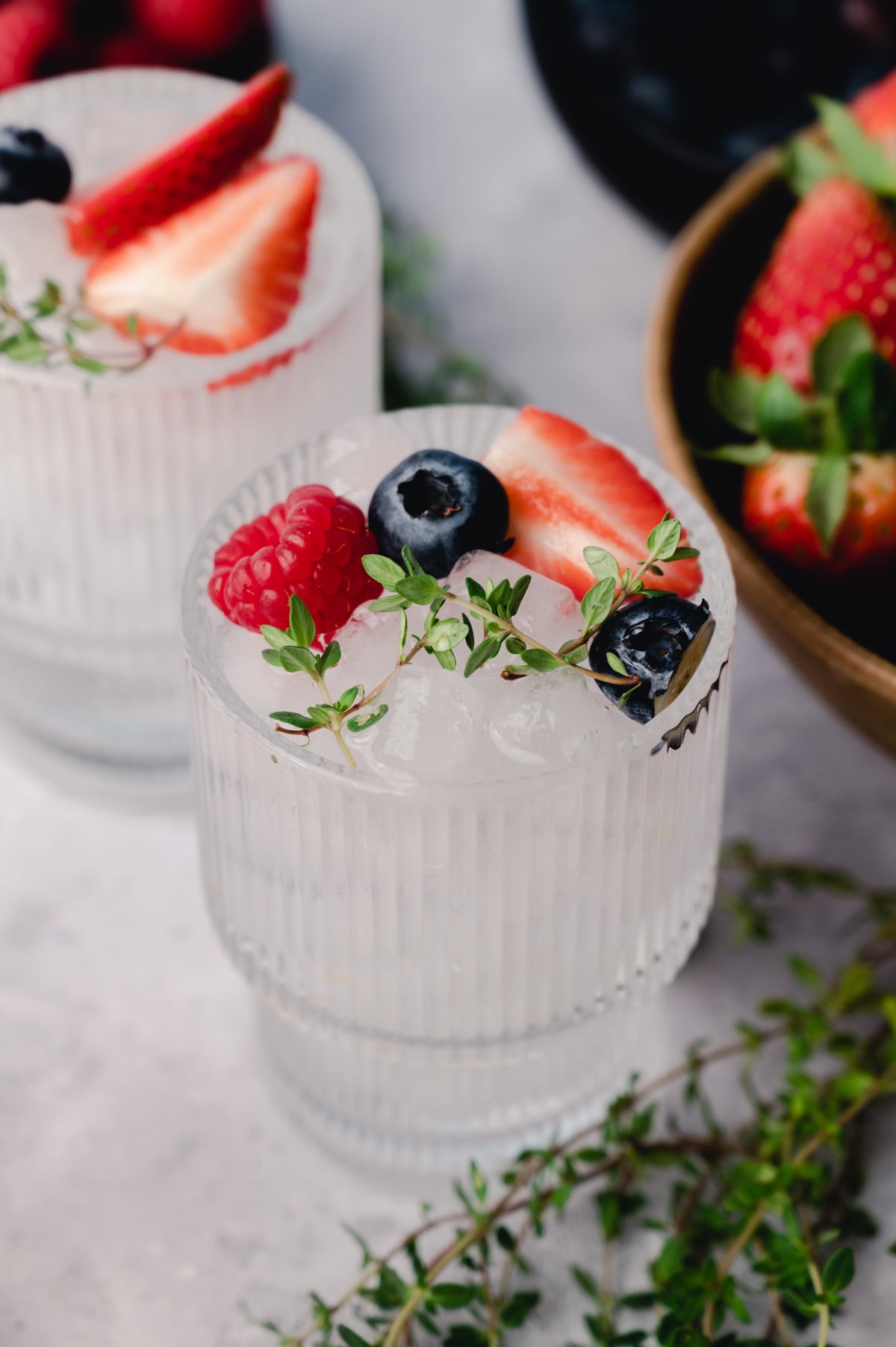 A clear glass filled with ice, garnished with a sliced strawberry, raspberry, blueberry, and a sprig of thyme. A bowl of fresh strawberries is in the background.