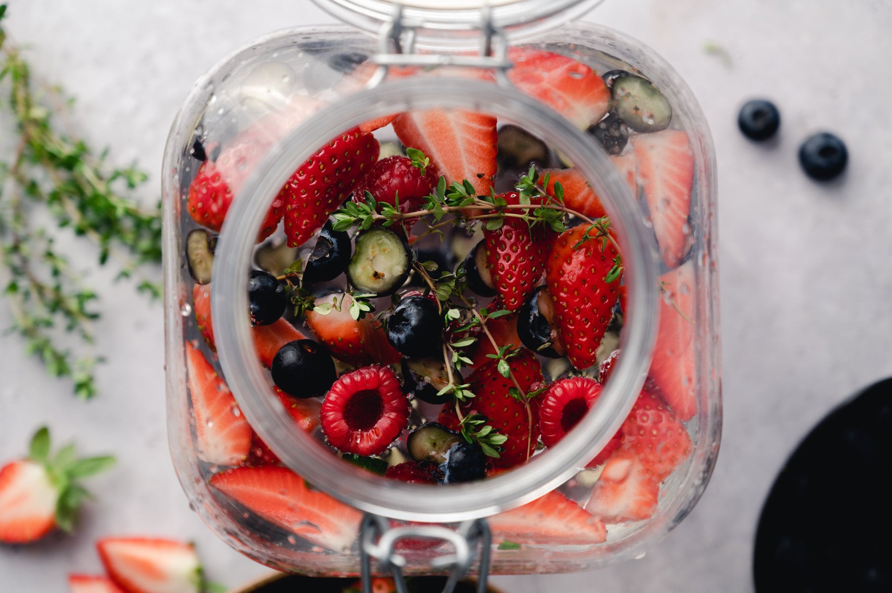 A glass jar filled with cut strawberries, blueberries, raspberries, and sprigs of thyme viewed from above. Some berries and thyme are scattered around the jar.