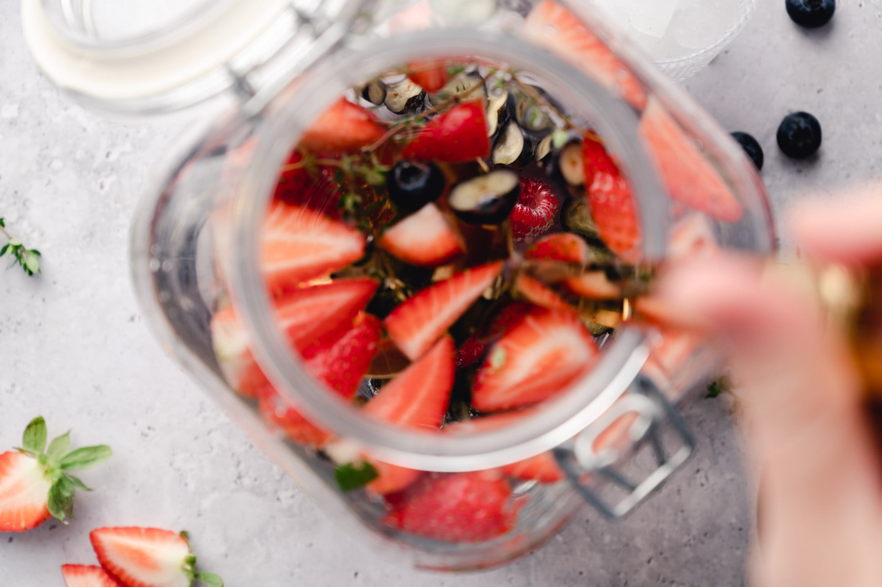 Top view of a jar filled with sliced strawberries and blueberries, placed on a light-colored surface. A person’s hand is visible, stirring the contents. Whole strawberries and blueberries lie nearby.