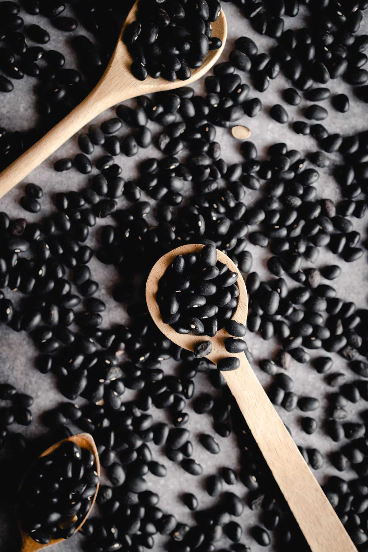 Three wooden spoons filled with black beans lie scattered among many loose black beans on a dark surface.