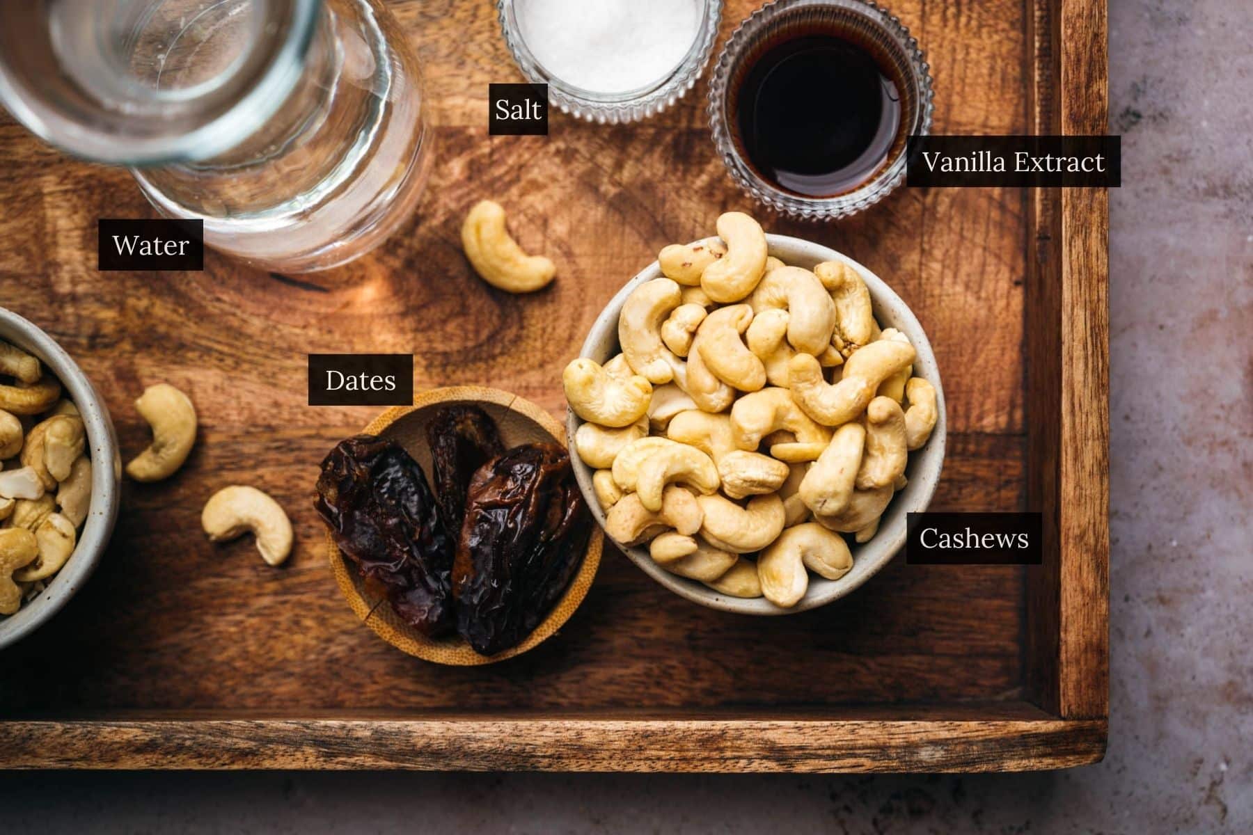 Wooden tray with bowls featuring cashews and dates, alongside a glass of water, labeled salt, vanilla extract.