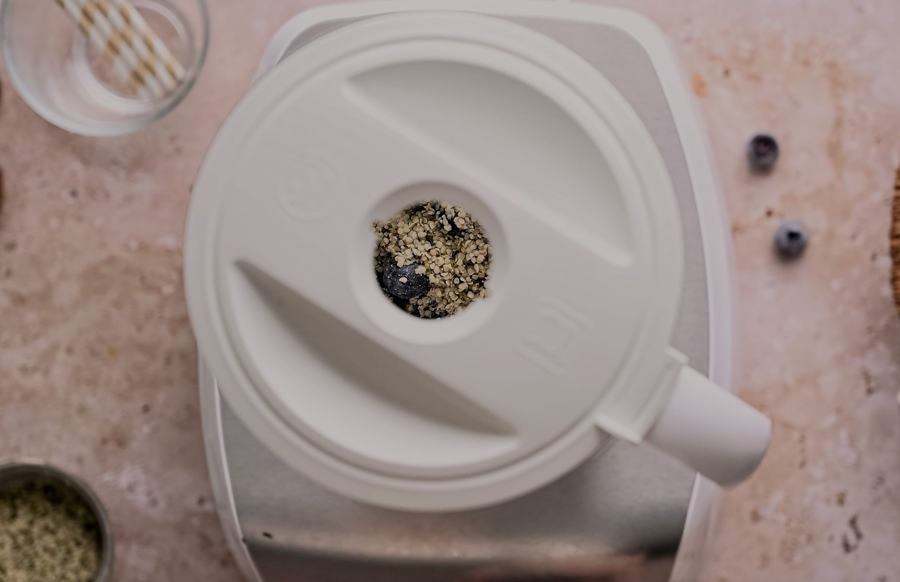 Top view of an open blender with a mixture of blueberries and seeds, surrounded by ingredients on a kitchen counter.