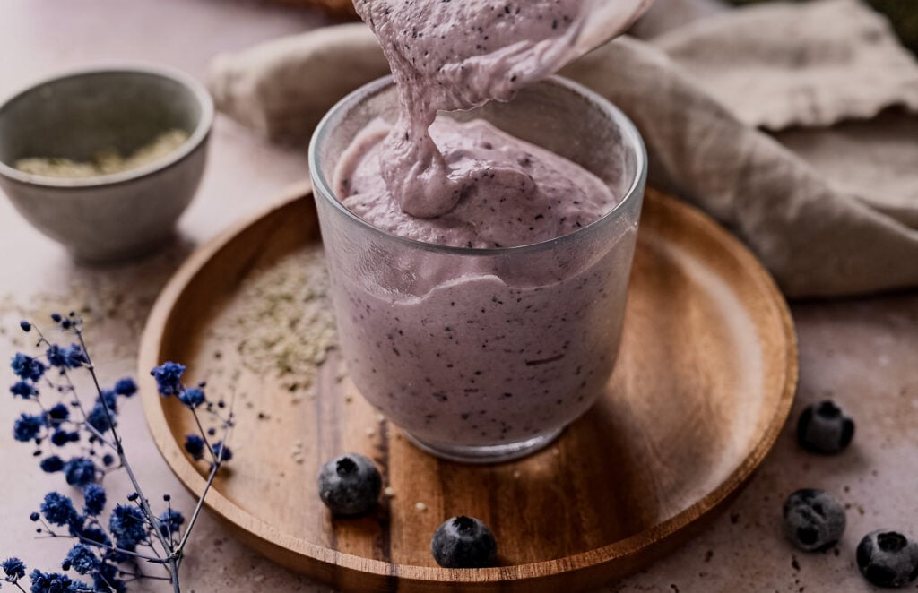 A creamy blueberry smoothie is poured into a glass on a wooden plate with fresh blueberries and a bowl of powder nearby.