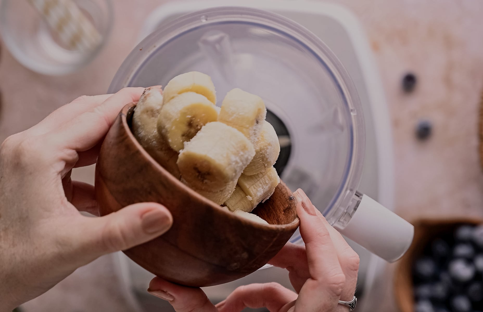 Hands holding a wooden bowl filled with sliced bananas, ready to be added into a blender with visible berries nearby.