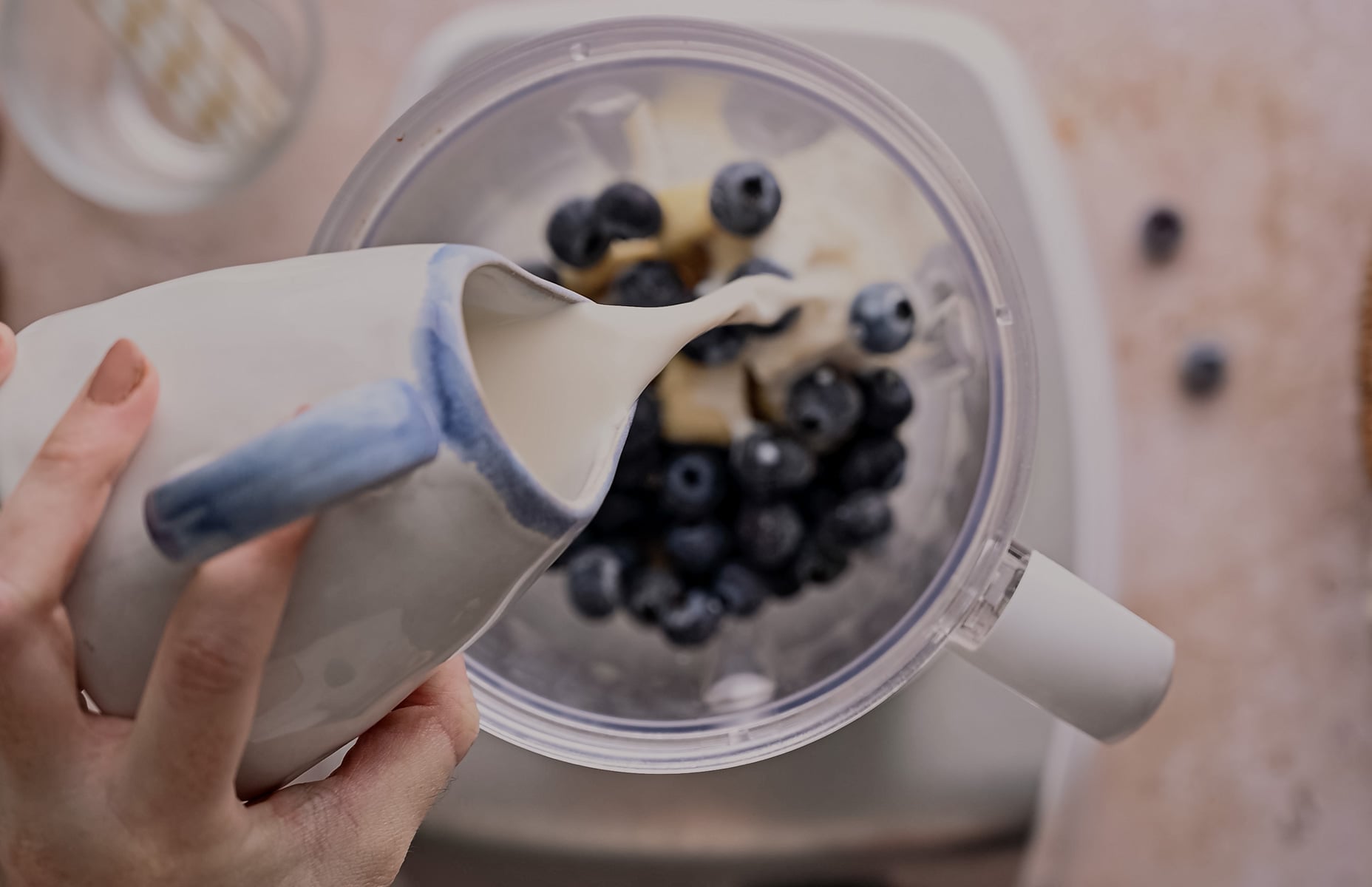 A person pours milk from a ceramic jug into a blender containing blueberries and bananas, with a marble countertop background.