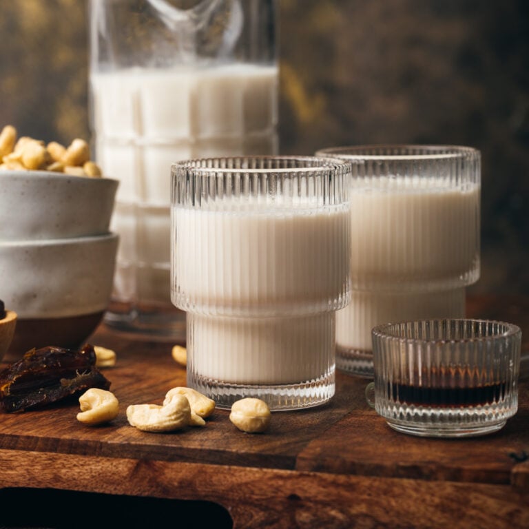 Two glasses of cashew milk, a small dish of liquid, a jug of milk, bowls of cashews, and dates are arranged on a wooden surface.