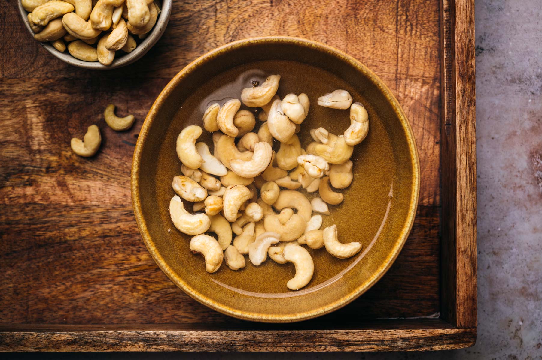A brown bowl filled with soaking cashew nuts is placed on a wooden tray, with a small bowl of dry cashew nuts beside it.