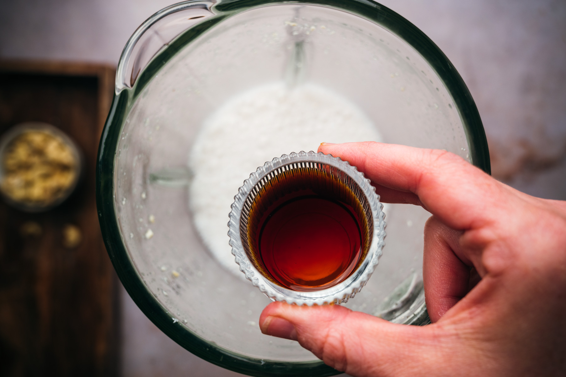 A hand pours a small glass of vanilla extract into a blender containing a white powder, possibly flour. There is a small bowl of nuts on a wooden surface in the background.