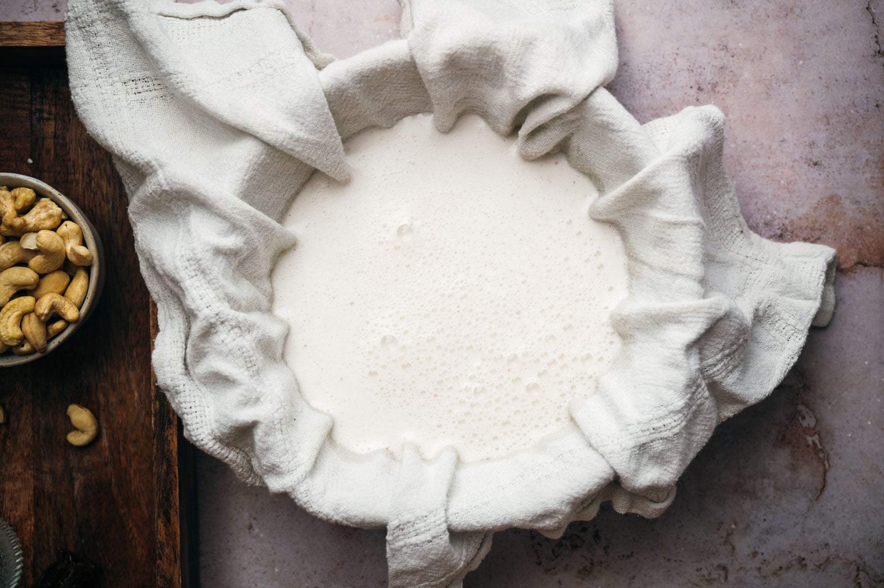 A bowl covered with a white cloth is filled with creamy liquid; cashew nuts are visible on a wooden surface nearby.