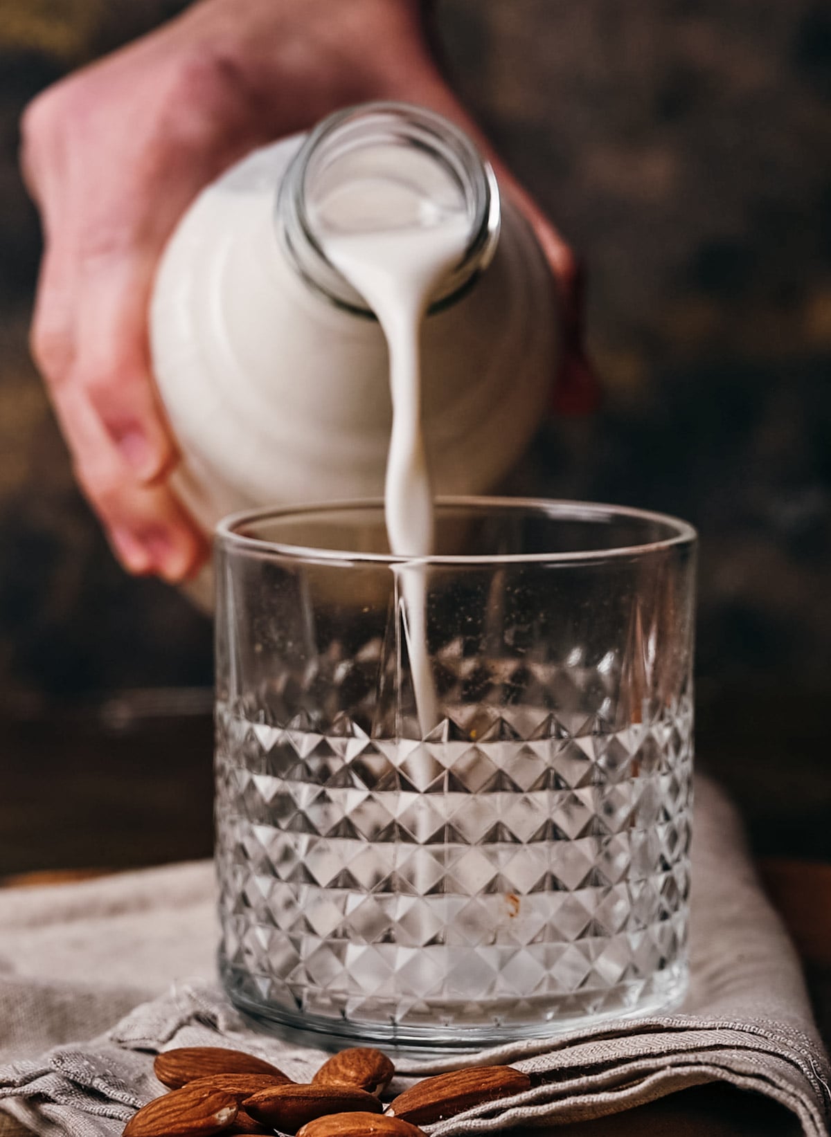 A person pours milk from a ceramic jug into a textured glass on a table with almonds scattered around.