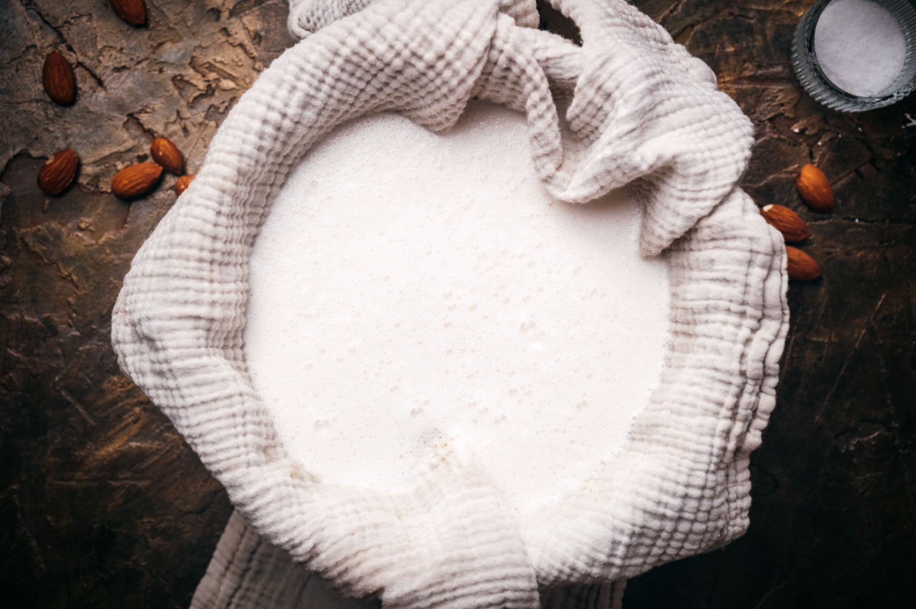 A top view of almond milk in a bowl wrapped with a white cloth on a rustic table, surrounded by scattered almonds.