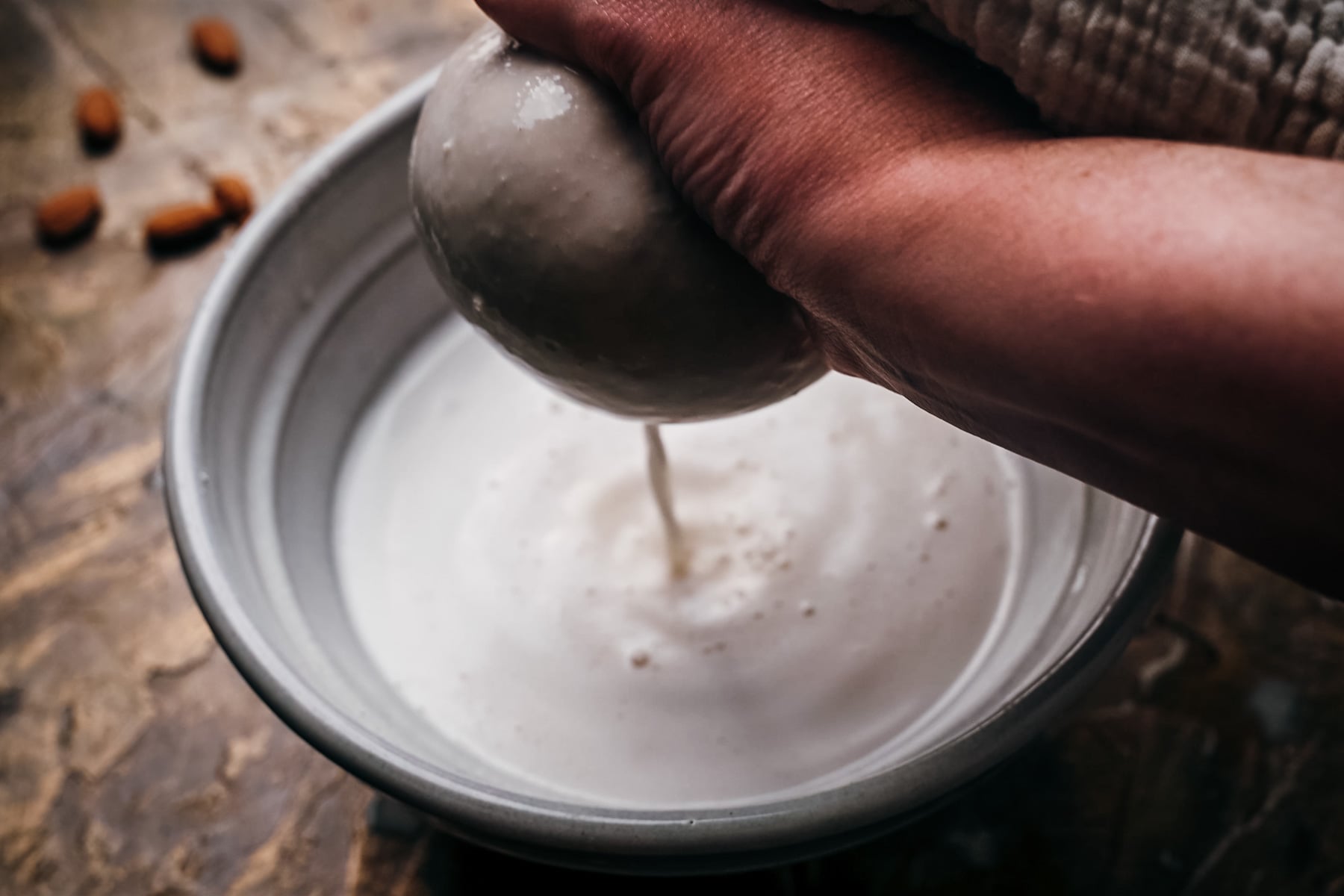 A person's hand is pouring almond milk from a cheesecloth into a bowl, with almonds scattered in the background.