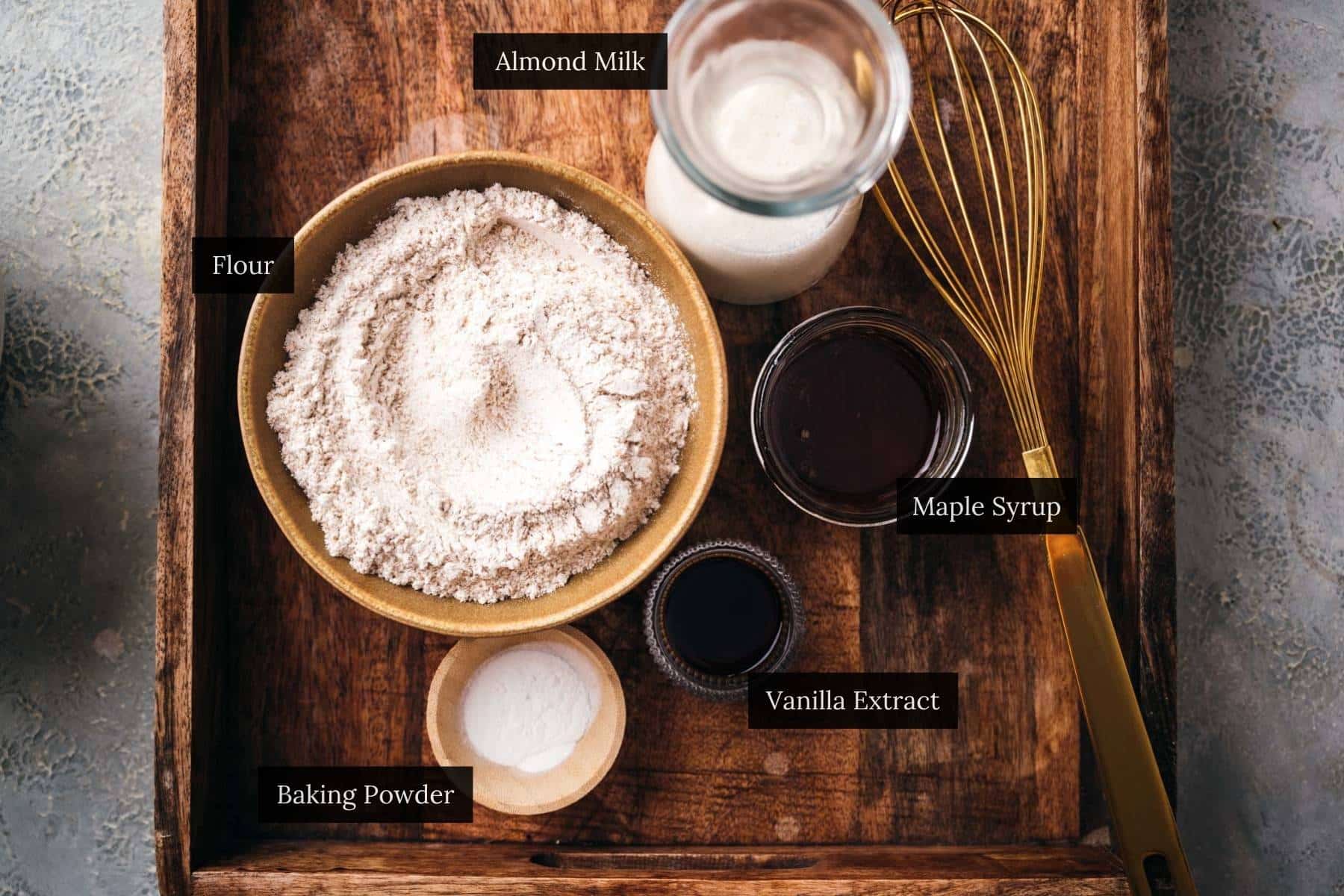 A wooden tray containing a bowl of flour, a bottle of milk, a whisk, a small bowl of baking soda, and two small jars filled with different dark liquids.