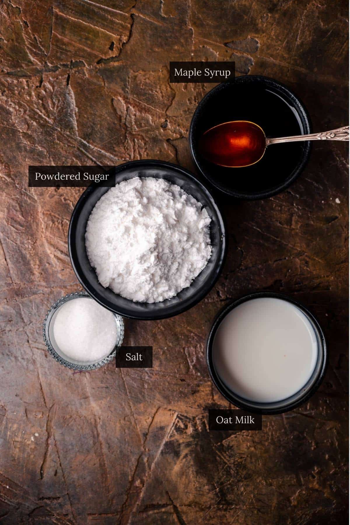 Powdered sugar, oat milk, and maple syrup in black bowl and salt in a class bowl. 