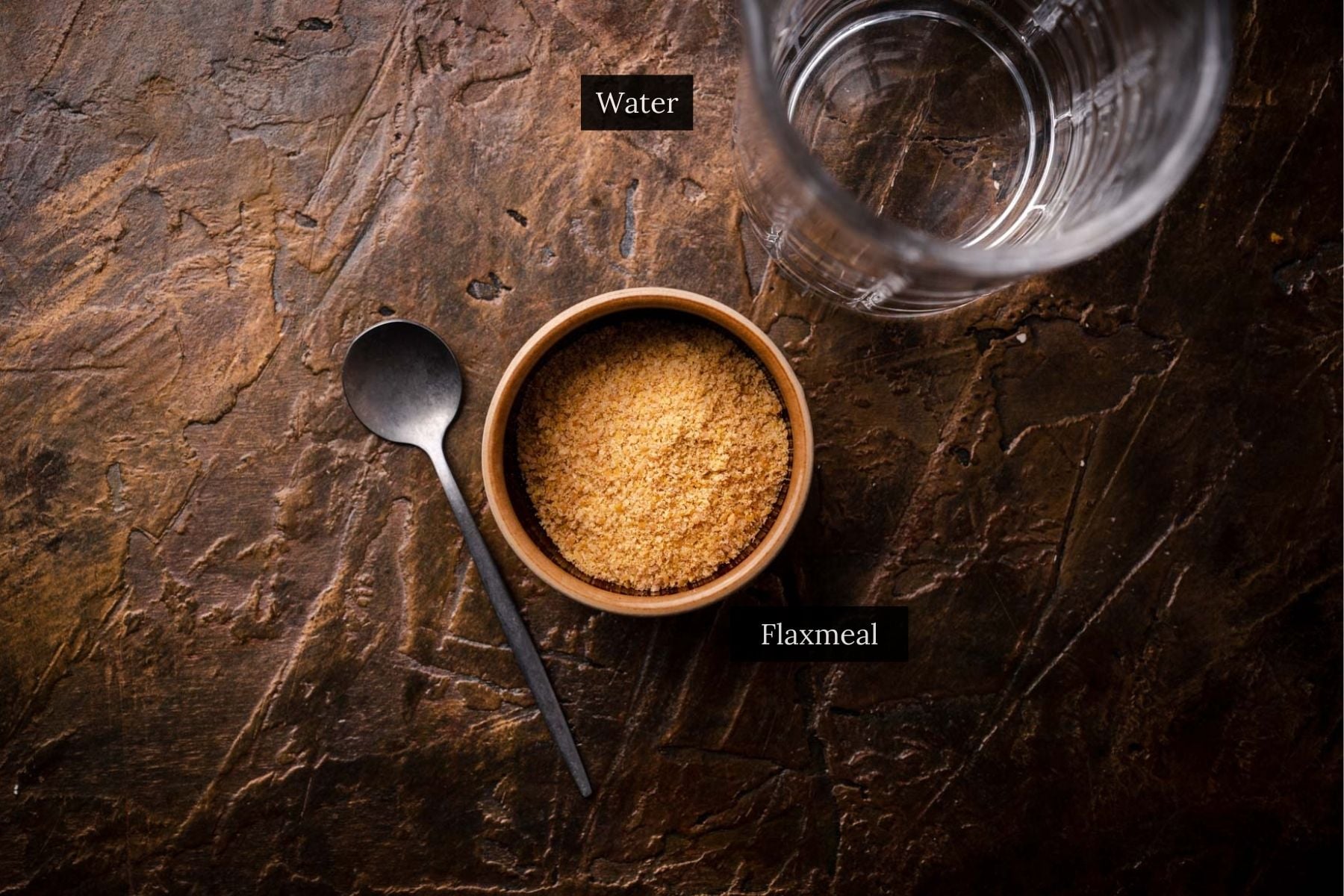 A glass of water, a bowl of flaxmeal for making a flax egg, and a spoon are arranged on a textured brown surface.