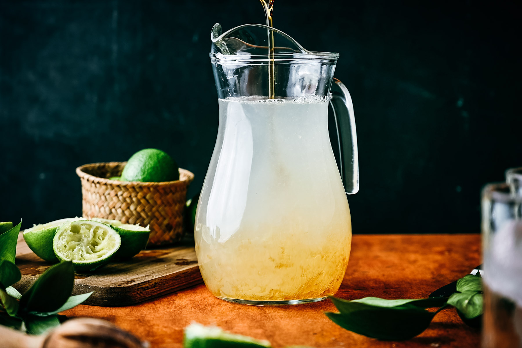 A pitcher of freshly made limeade on a wooden table, with cut limes and a basket nearby, against a dark background.