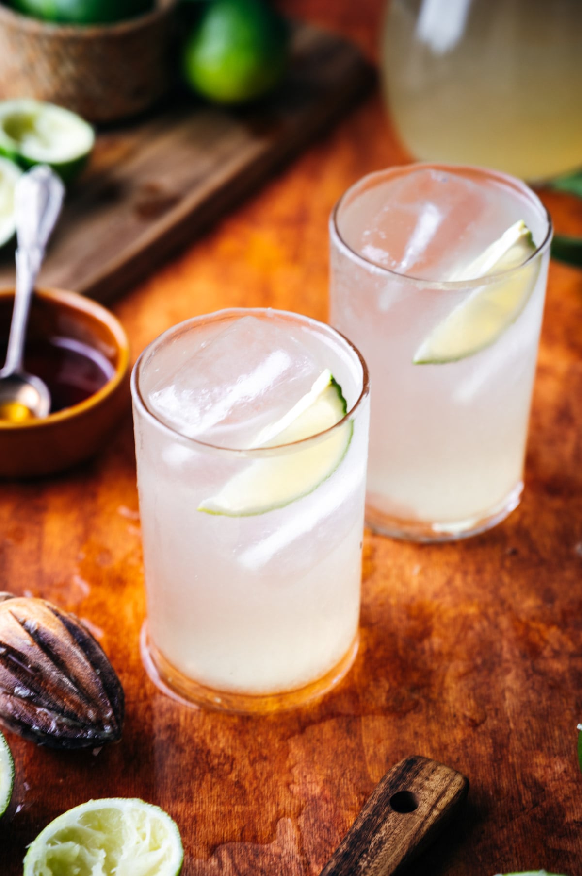 Two glasses of limeade with ice and lime slices, on a wooden table surrounded by halved limes and a rustic background.