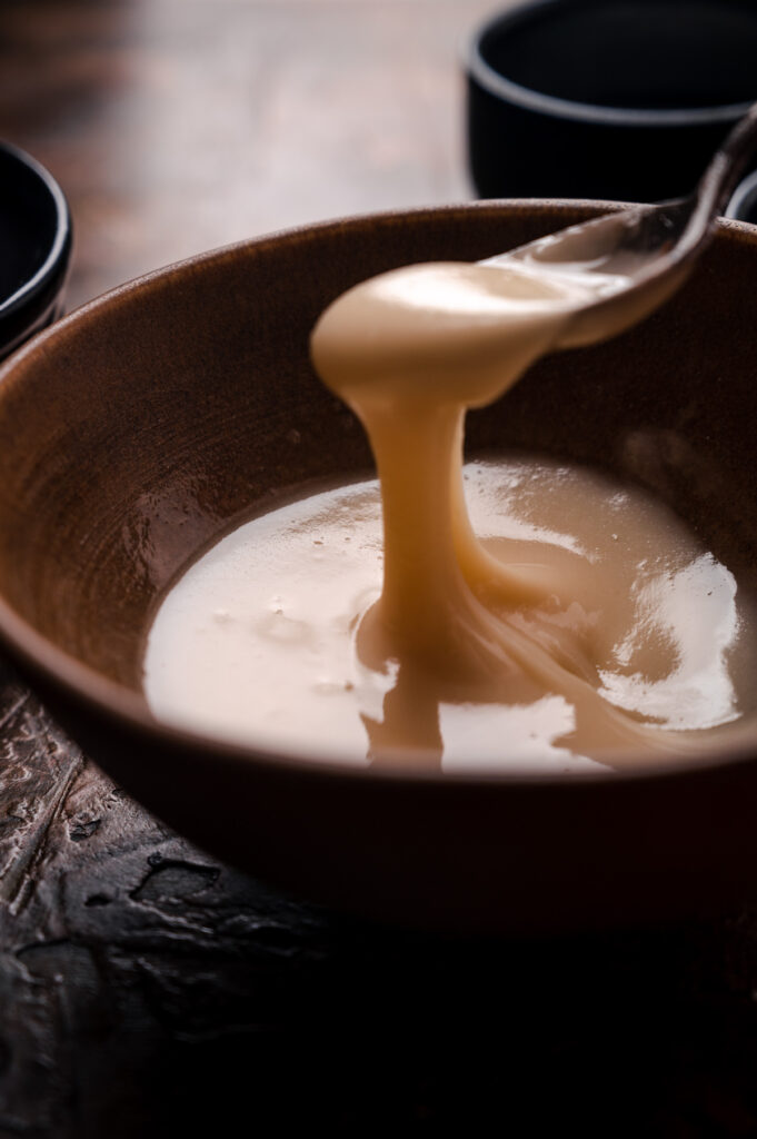 A close-up image of a spoon drizzling thick, maple glaze into a rustic brown bowl on a wooden table.