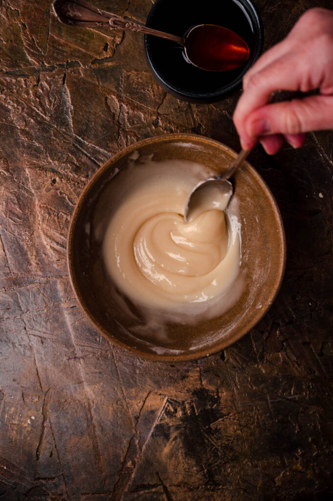 A hand stirring a thick, creamy sauce in a rustic bowl with a spoon, placed on a textured dark wooden surface with a cup of sauce nearby.
