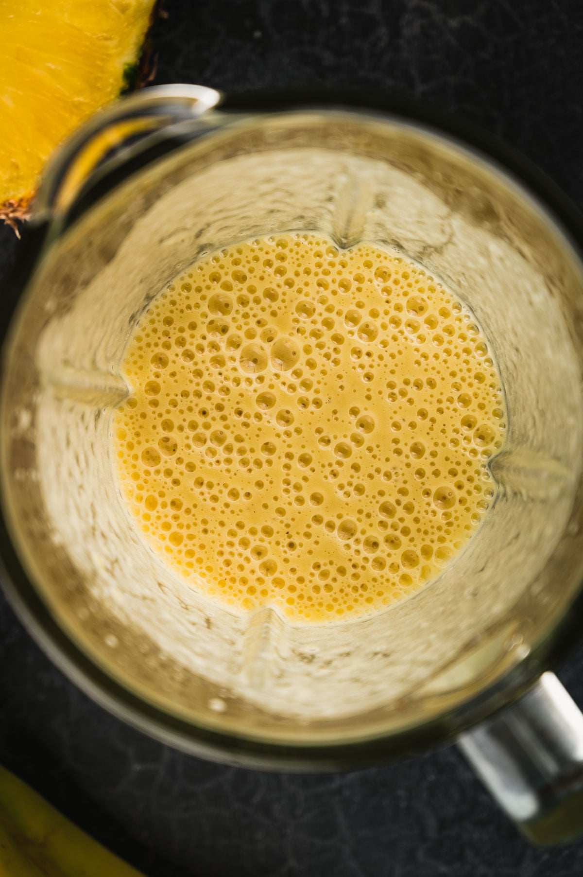 A close-up view of a blender containing a yellow, frothy smoothie mixture. The blender is placed on a dark surface with a hint of a pineapple visible at the top left corner.