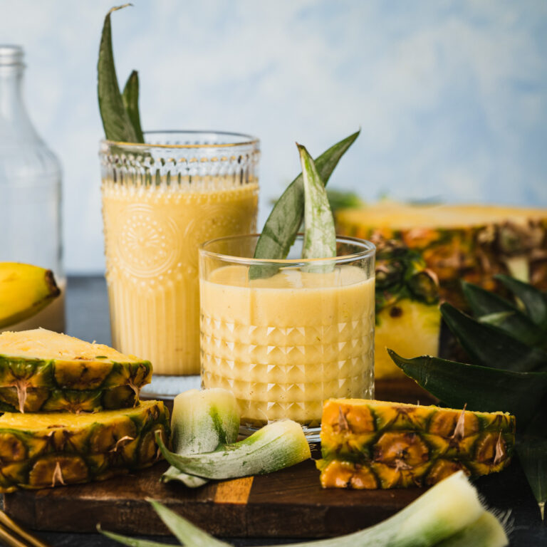 Two glasses of fresh pineapple smoothie, one with a decorative pattern, surrounded by sliced pineapples and pineapple leaves on a wooden surface. A whole pineapple and a banana are in the background.